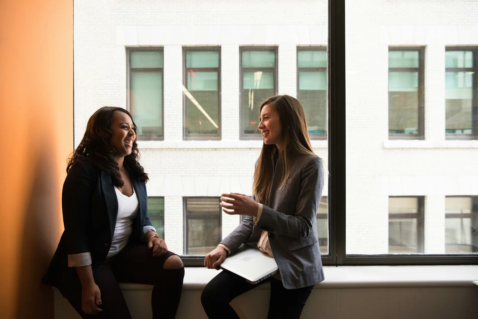 Two women laughing in conversation at a table by a window